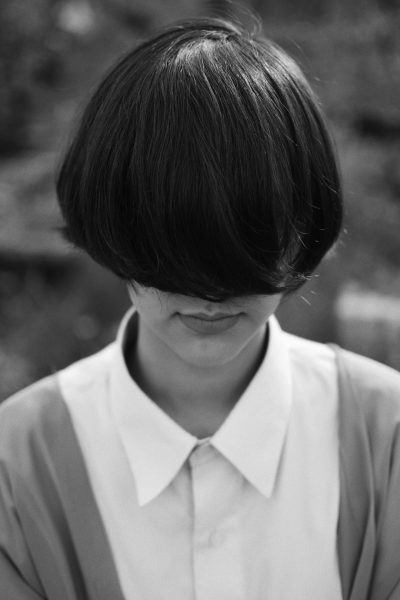 A black and white photo of an Asian woman with short hair wearing a shirt, looking down at the ground with her head tilted to one side. She has bangs that cover half or more of her face. The background is blurred, creating soft focus on her face. Shot in the style of Tadao C, she stands in front of some trees. Her expression appears sad but still confident as if it were something she had faced before. --ar 85:128