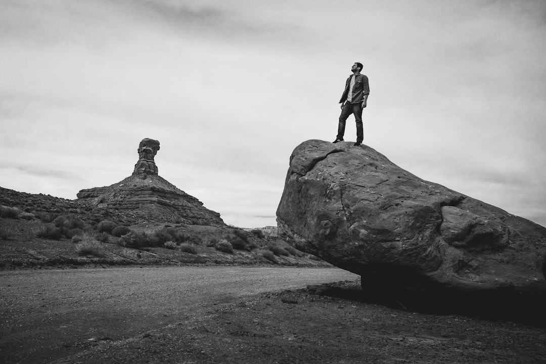 Black and white photography of an man standing on top of rocks in Moab, Utah during the day with natural light. The rock formations are unique shapes. There is a road leading to them. He wears stylish business attire. –ar 128:85