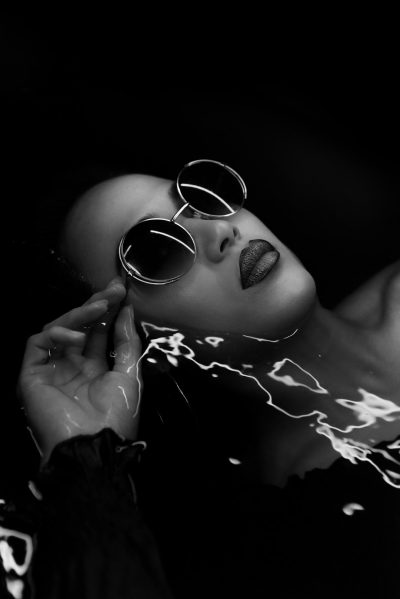 A black and white studio portrait of a bald woman with silver sunglasses floating in water, the glasses have light coming out from them creating lightning-like structures around her face. She has beautiful makeup on, her hair is wet and long. The background is plain black, the lighting creates deep shadows that accentuate her features, creating dramatic contrast between dark tones and bright highlights in the style of [Rembrandt](https://goo.gl/search?artist%20Rembrandt). --ar 85:128