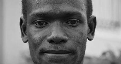 A close-up portrait of an African man in the streets, photographed with a Fujifilm XH2S and Fujinon XF lens, in black and white photography in the style of grainy film noir. --ar 32:17