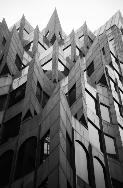 A black and white photo of the facade of an office building in downtown Boston in the style of [Frank Gehry](https://goo.gl/search?artist%20Frank%20Gehry) with angular, symmetrical geometry that resembles concrete vertical triangles or diamonds. --ar 83:128