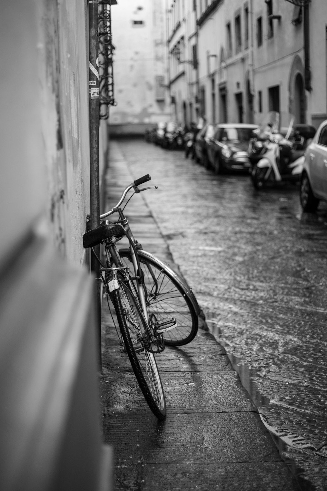 Black and white photography of an old bicycle leaning against the wall in a narrow street, surrounded by parked cars, on wet streets of Firenze during rainy day. –ar 85:128