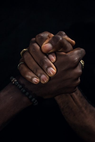 A closeup of two hands, one black and the other white, clasped in prayer against an all-black background, symbolizing unity during times like Black history month. The focus is on their interlocked fingers with detailed textures of skin color, rings, bracelets, showcasing diversity and contemplation through shared religious practice. This portrait captures a moment of connection between individuals from different ethnic backgrounds in the style of religious artwork. --ar 85:128