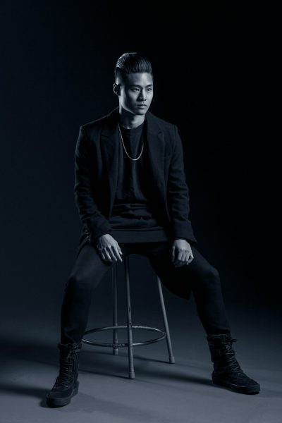 full body photography of an Asian man sitting on a bar stool, wearing a black blazer and t-shirt with dark pants, clean shaven face, very short hair style, posing for a photoshoot, dark background, studio lighting, black and white. --ar 85:128