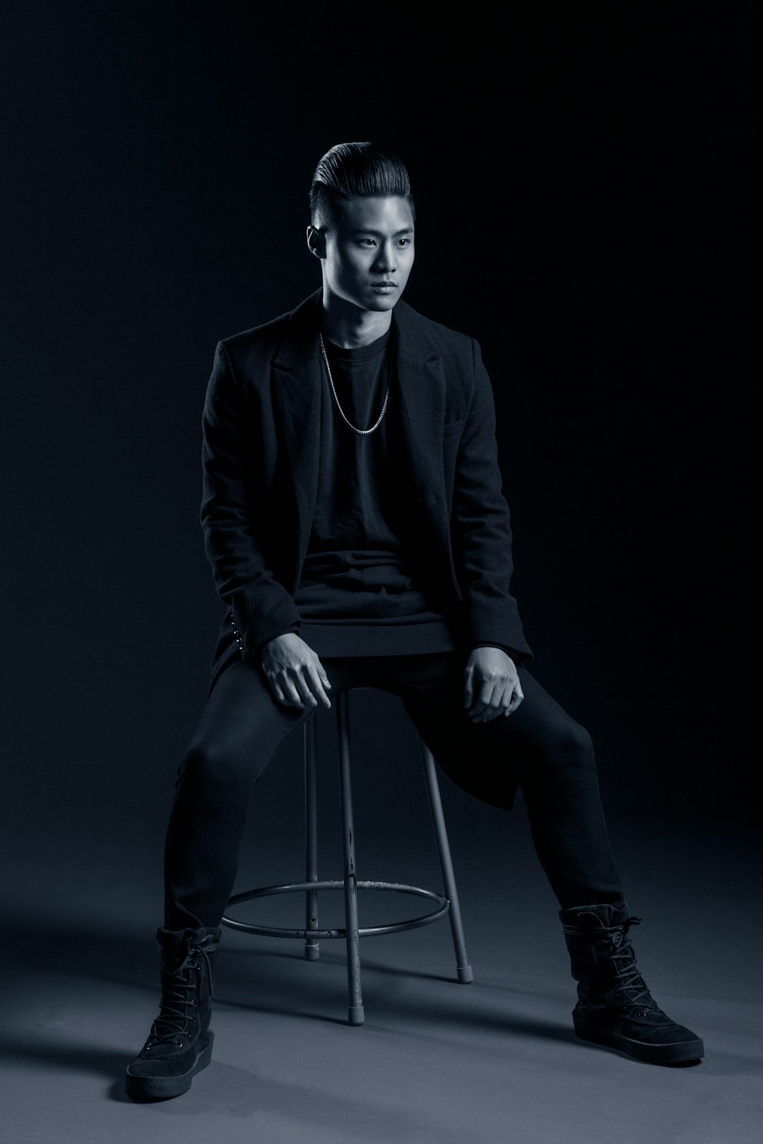 full body photography of an Asian man sitting on a bar stool, wearing a black blazer and t-shirt with dark pants, clean shaven face, very short hair style, posing for a photoshoot, dark background, studio lighting, black and white. –ar 85:128