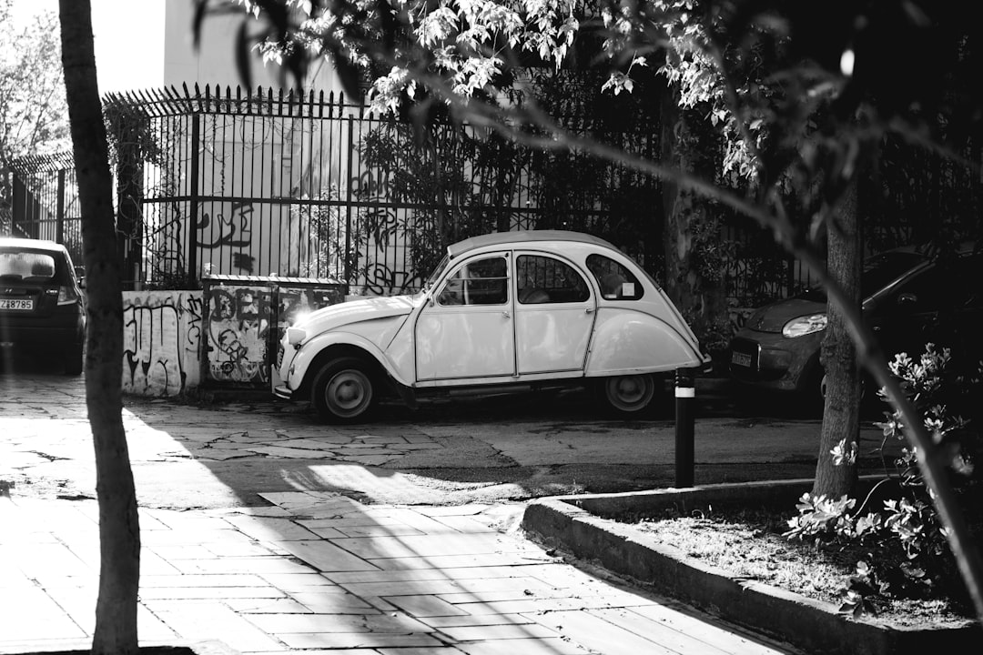 Black and white street photography of an old white Citroen car parked in the streets of Lille, France. The shot is taken from across the road, and shows part of some trees and shrubs to give context as well as other cars on the side street. It’s late afternoon with soft light. –ar 128:85