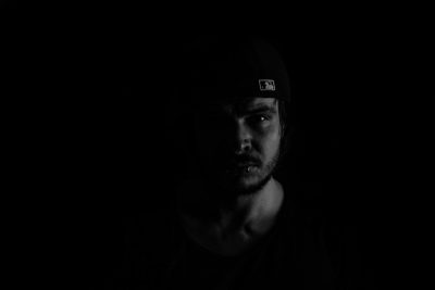 Black background, man wearing cap with white logo on it, looking at camera, dark portrait, minimalism, black and white, low light photography, blurred edges, dark tones, symmetrical composition, halflength shot, clear facial features. --ar 128:85