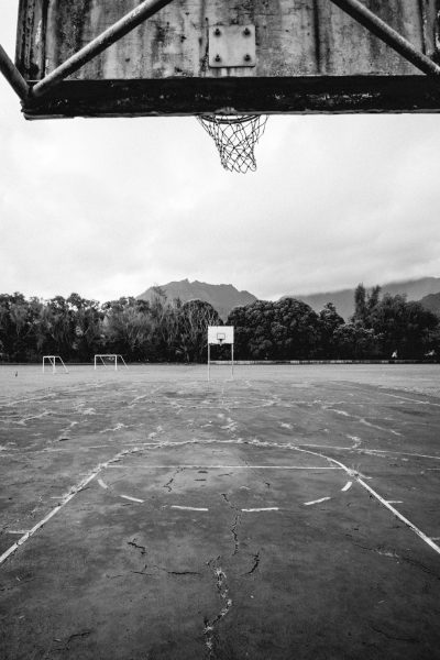 A wide-angle view of an abandoned soccer field with concrete floors, the goal frame is visible in front and behind it there is a forested mountain range. The photo was taken from under one basketball hoop looking up at another, the sky above is overcast. Black and white photography, shot in the style of Kodak Gold 400 film stock. --ar 85:128