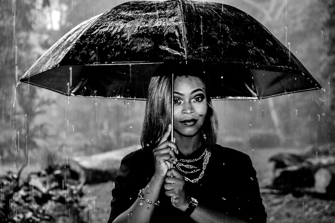 African American woman in her mid-30s, wearing black and white attire with jewelry accents, standing under an umbrella on a rainy day, portrait photography with a Nikon D850, capturing the raindrops and wet hair texture in a black & white photograph with high contrast between the subject’s skin color and background and soft lighting emphasizing facial expression. –ar 128:85