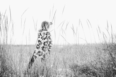 A black and white photograph of a young girl wearing a flower pattern coat, standing in tall grass near the beach on Long Island. The photograph has a minimalist, high contrast style with the back view of the girl looking out to the sea. The photograph was taken with a Fujifilm XH2S camera. --ar 128:85