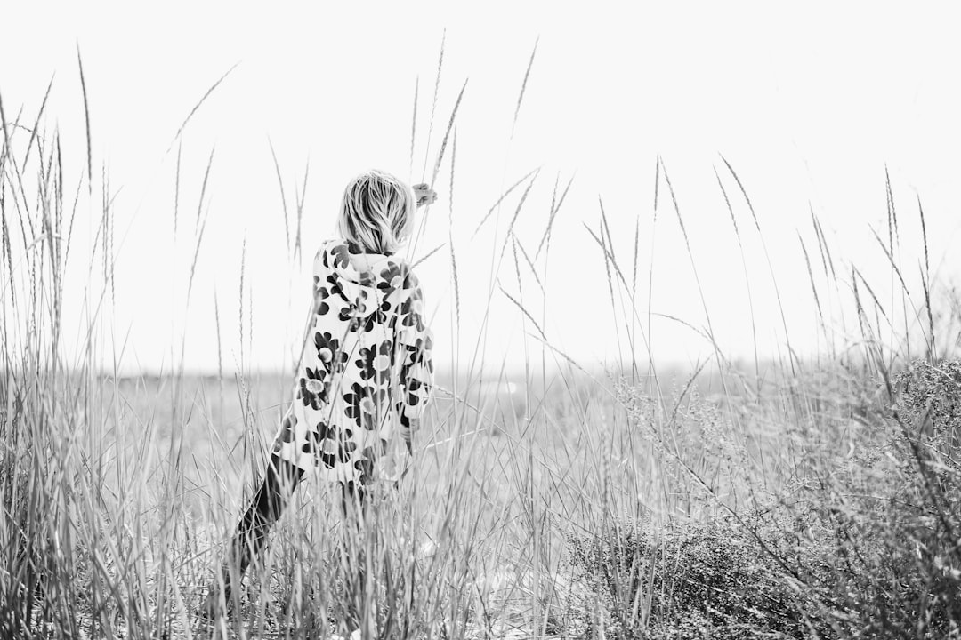 A black and white photograph of a young girl wearing a flower pattern coat, standing in tall grass near the beach on Long Island. The photograph has a minimalist, high contrast style with the back view of the girl looking out to the sea. The photograph was taken with a Fujifilm XH2S camera. –ar 128:85