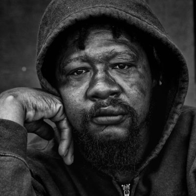 Black and white close up portrait of an African American homeless man wearing a hoodie, looking at the camera with one hand on his cheek, in downtown Los Angeles. In the style of Leica M6.