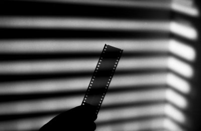 A hand holding up an old film strip, with the shadow of blinds in the background. The film reel is slightly blurred and has small white dots on it. Black and White Film photography. Shot taken by Sony Alpha A7 III camera with a extreme long shot angle. --ar 128:83