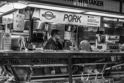 Black and white street photography of an unidentifiable man working at the counter inside his pork chip stand, with large signs above him that say "PMombo" in bold letters on top of each other. There is also another sign below saying it's for carved out or wrapped pig pure eye pale pink ham strips called "CAmbre CUst", "V marty Bages". The scene includes an open kitchen behind where he makes Han from fresh raw meat to runny chrisps, and lots more food items such as bags of chips and pop, --ar 128:85