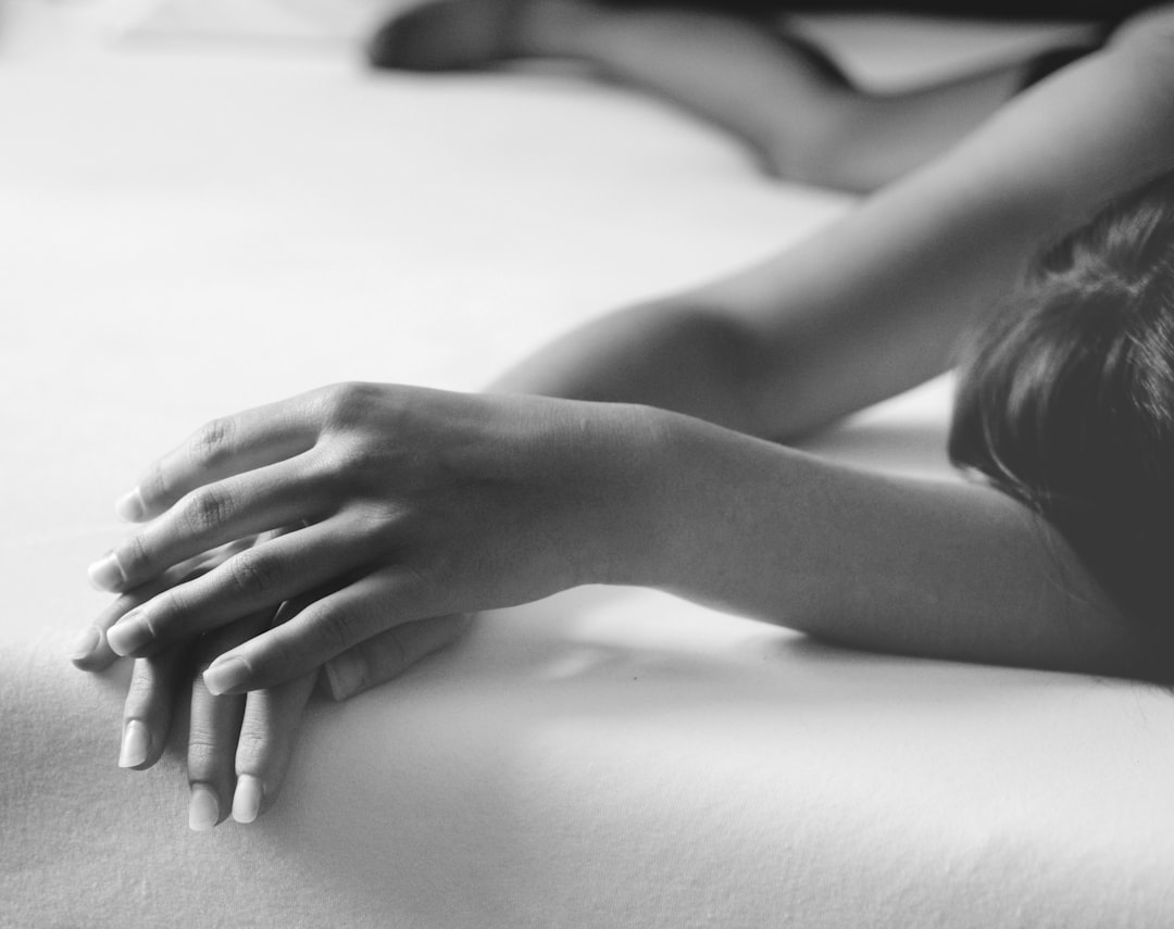 A black and white closeup photograph of the backside, hands holding her waist on top, showing only one hand in focus. The woman’s skin is smooth with soft lighting that highlights its texture. She has long hair tied behind her. Her fingers are adorned with elegant nail polish. In front view, she lies flat down on an empty bed, creating a sense of calmness. This composition creates a serene atmosphere, focusing attention to both beauty and tranquility. –ar 128:101