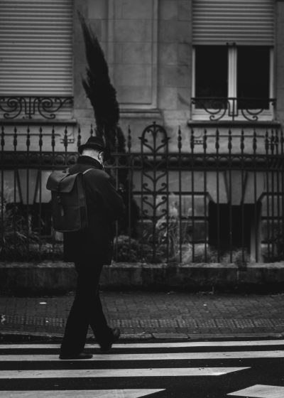 A man in a suit and tie with a backpack crossing the street on zebra stripes, Parisian architecture, a black metal fence, film grain, monochrome photography, in the style of Leica M6, 35mm f/4 lens, f/20 aperture, vintage filter. --ar 91:128