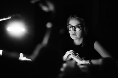 Black and white photography of an influencer being interviewed in the style of the photographer, illuminated in a dark room with soft lighting. The photo is taken from behind her as she sits at a desk. She has blonde hair and glasses. Her face looks directly into the camera. There is another person sitting next to her who you can't see clearly. --ar 128:85