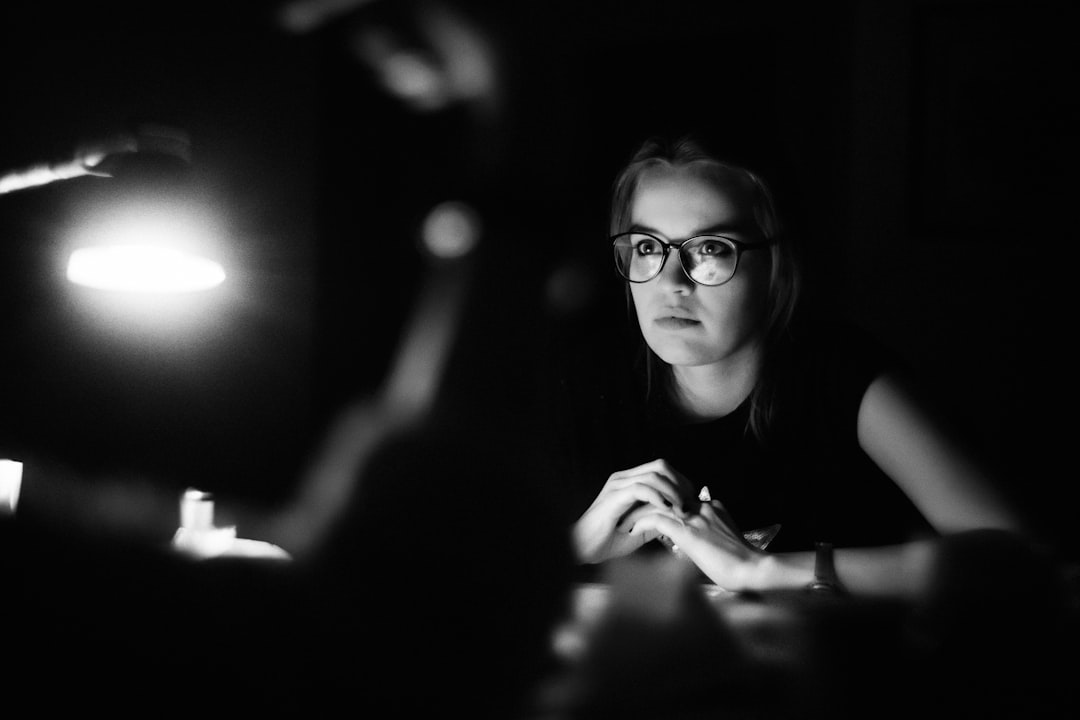 Black and white photography of an influencer being interviewed in the style of the photographer, illuminated in a dark room with soft lighting. The photo is taken from behind her as she sits at a desk. She has blonde hair and glasses. Her face looks directly into the camera. There is another person sitting next to her who you can’t see clearly. –ar 128:85