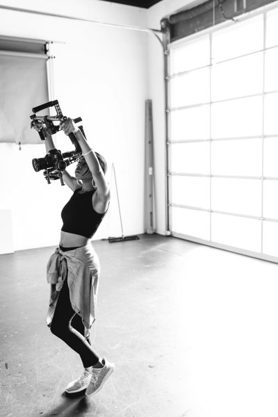 A black and white photo of an influencer carrying her camera on her shoulder, wearing sportswear in front of large garage doors. She is dancing with joy while holding up one hand to film the dance scene. The floor nearby has studio lighting equipment scattered around it. In a modern minimalist style, this shot captures a moment frozen, showcasing elegance through dynamic movement and expressive facial expressions in the style of the lens. --ar 85:128