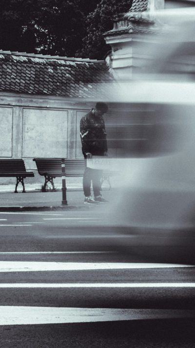 A blurry black and white photo shows two people in the street, one standing on an empty bench with his back to the camera while another person passes him. The focus is slightly blurred around them, capturing motion blur. They both wear sneakers. In front is a pedestrian crossing with visible stripes. There are also some other elements like buildings or trees in the background. The shot was taken from behind, the scene appears very minimalistic and simple in the style of a minimalist photographer. --ar 9:16