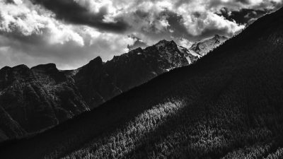 black and white photo of the rocky mountains, the mountain range, black clouds, dark sky, mountain side, forest, sun rays, shadows, shot with a Sony Alpha A7 III camera, using a Sony FE 24-85mm f/3.0-4.5 lens, with sharp focus, high resolution, capturing global illumination, using natural light, in the style of professional photography, using HDR, with very detailed imagery. --ar 16:9