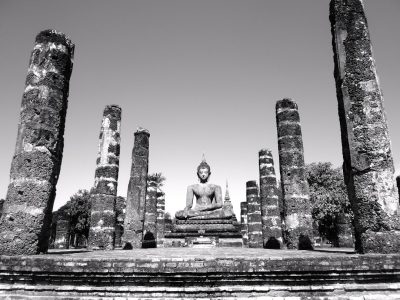 Black and white photography of an ancient temple with broken pillars, the main focus is on a sitting Buddha statue in front of it. The scene captures the grandeur of the old architecture against clear skies. --ar 4:3