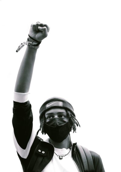 A black and white photo shows an African American man wearing streetwear. He has one arm raised high in the air, holding up his fist. A black bandana covers his head and a face mask. He has dreadlocks. The man is isolated against a solid white background. The photo is shot from above, looking down at him as he raises his hand towards the sky. The style of the photo resembles that of a famous artist. --ar 45:64