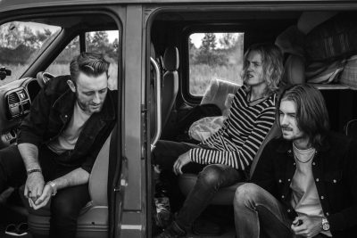 A black and white photo shows three men sitting in the back doors of an open van. One man wears a striped shirt with long hair and facial stubble. The other two have short dark blonde hairstyles and look happy as they play music together inside the vehicle. They all wear leather jackets. In front, another guy has his hands on his knees looking at them. He wears glasses and looks like a young rockstar. It’s a sunny day. Shot in the style of [David LaChapelle](https://goo.gl/search?artist%20David%20LaChapelle). --ar 128:85