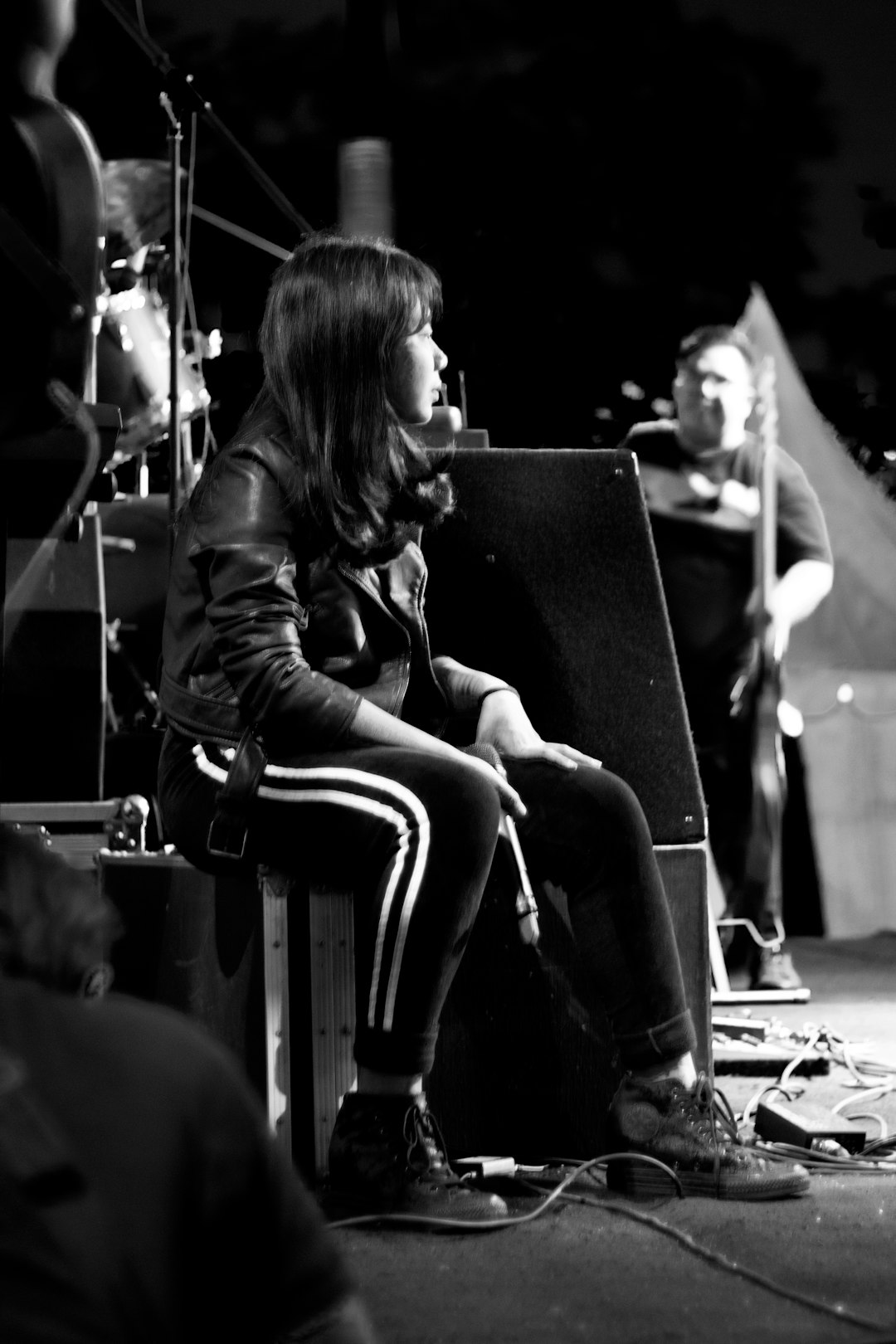 A black and white candid photo of an alternative woman with shoulder-length hair wearing a leather jacket, striped pants and Converse shoes sitting on top of a stage arm chair in front of her musical band during magic hour, with film grain, in a full body shot, captured on Fujifilm Pro 400H film in the style of a candid photo. –ar 85:128