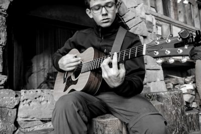 A young man with glasses playing acoustic guitar, sitting on stone steps outside of an old cabin in the woods, in the style of black and white photography. --ar 128:85