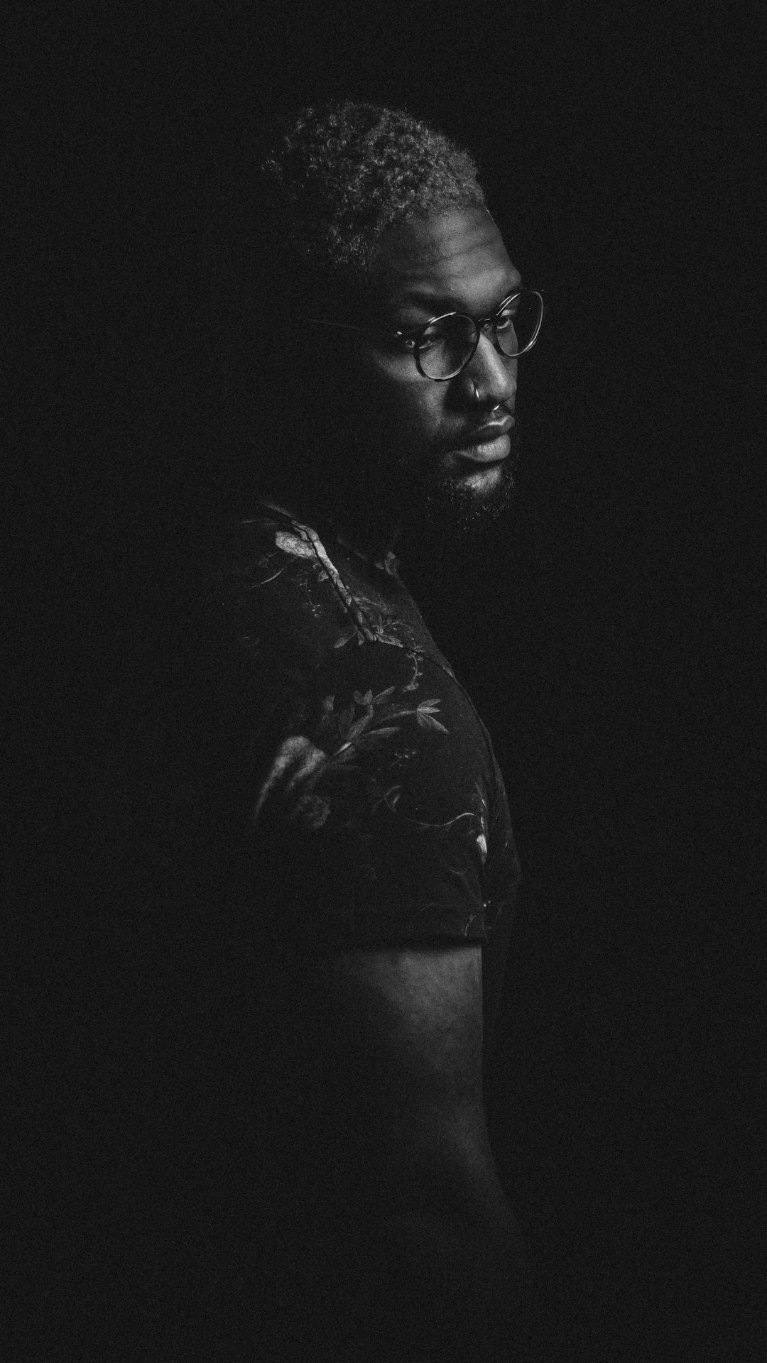 A black and white portrait of an African American man with glasses, standing in the dark, wearing streetwear . He has short curly hair and a beard, illuminated by soft side lighting that casts dramatic shadows on his face and body. The background is completely black, emphasizing his silhouette against the light from behind. He’s looking slightly to one side, adding depth to the composition. Shot with a Leica M6 using Ilford HP5 film stock in the style of street photography. –ar 9:16