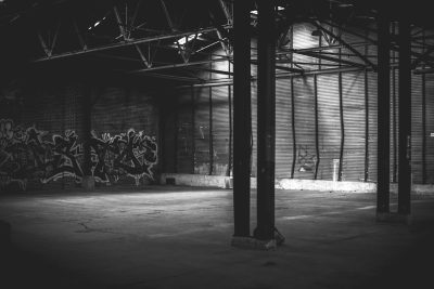 An empty warehouse with graffiti on the walls, captured in black and white photographic style. The scene is illuminated by soft light that casts gentle shadows across its vast expanse. A few metal columns stand tall against one of the brick walls, adding to the industrial feel of the space. This photograph captures an atmosphere filled with raw energy and urban coolness in the style of [Banksy](https://goo.gl/search?artist%20Banksy). --ar 128:85