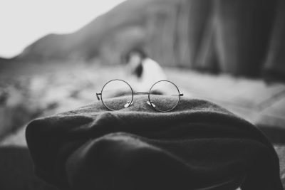 Black and white photo of round glasses resting on the edge of an old blanket, with blurred mountains in the background. The scene is captured in the style of behind the glasses. --ar 128:85