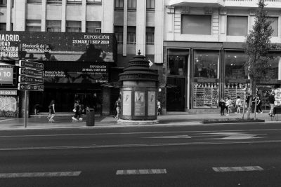 A black and white photo of the street corner in Madrid with an outdoor exhibition stand for "Mprobably based on S fertility" that is visible from across the road, showing people visiting it. The sign reads 'Ex veto 23 tats de G pseudo bryophones y m building' A public toilet stands next to them. In front there's some advertising boards for stores nearby, a man wears casual walking around, in style raw. --ar 128:85