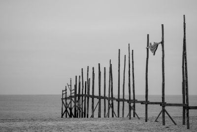 A black and white photograph of an old wooden pier with many tall, thin vertical planks standing on the beach in front of it. A small piece of fabric is hanging from one post. The sea can be seen at its edge. There are no people or animals visible. In the style of documentary photography. --ar 128:85