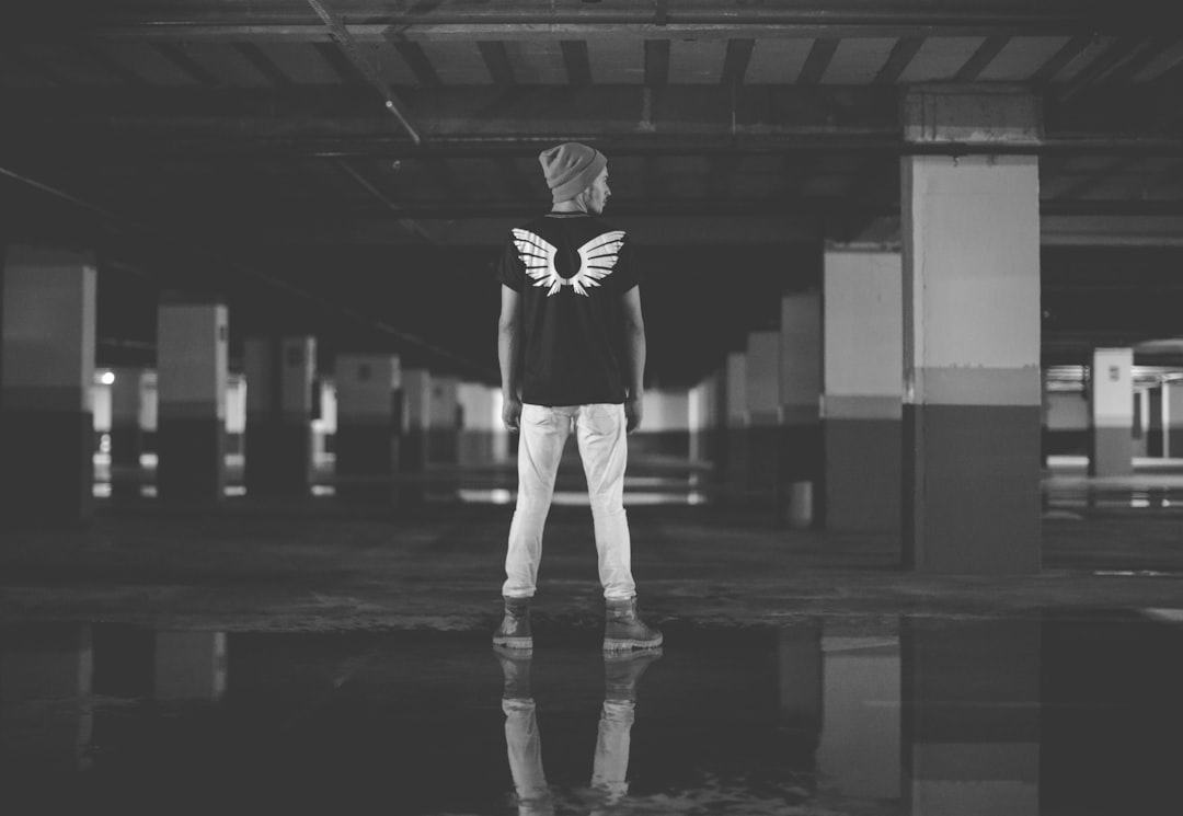 A black and white photo of an angel wearing jeans, standing in the middle of empty parking garage, wearing tshirt with wings on it, he is facing away from camera, full body shot, unsplash photography style, minimalistic –ar 16:11