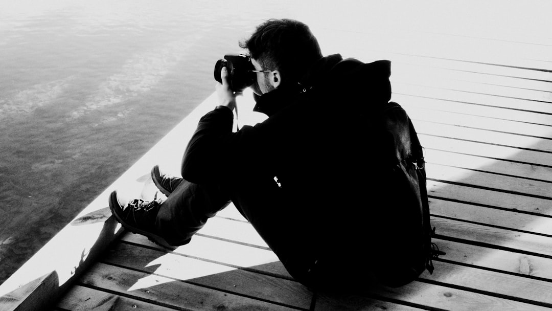 Black and white photo of a photographer sitting on the edge of a wooden dock, holding a camera to his eye while taking pictures of a lake. In a black jacket with a backpack on his back. –ar 16:9