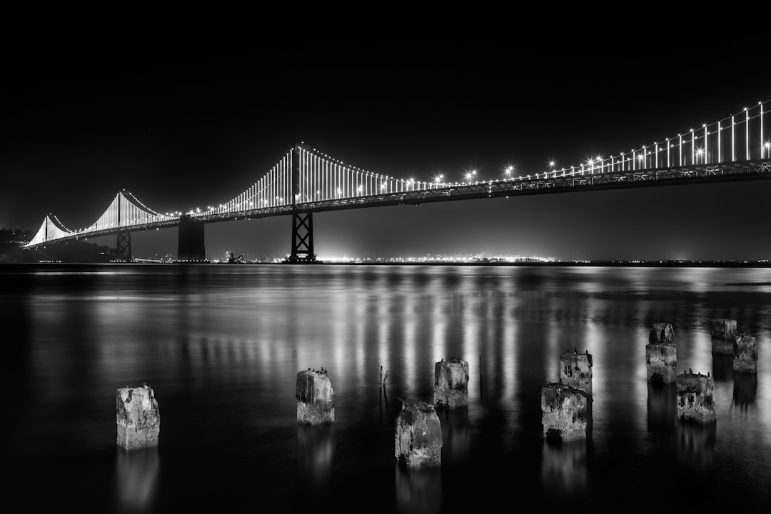 A black and white photo of the Bay Bridge at night with wooden posts in front of it, reflection on the water, wide shot, cinematic in the style of [Ansel Adams](https://goo.gl/search?artist%20Ansel%20Adams). –ar 128:85