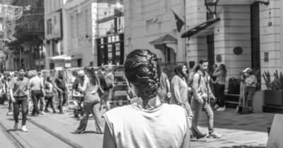 A young woman with her hair in an elegant French braid stands on the street of Milan, surrounded by people who walk along it and look at her. The photo is in the style of black and white street photography, taken from behind the subject with a back view using a Canon EOS R5 camera. --ar 128:67