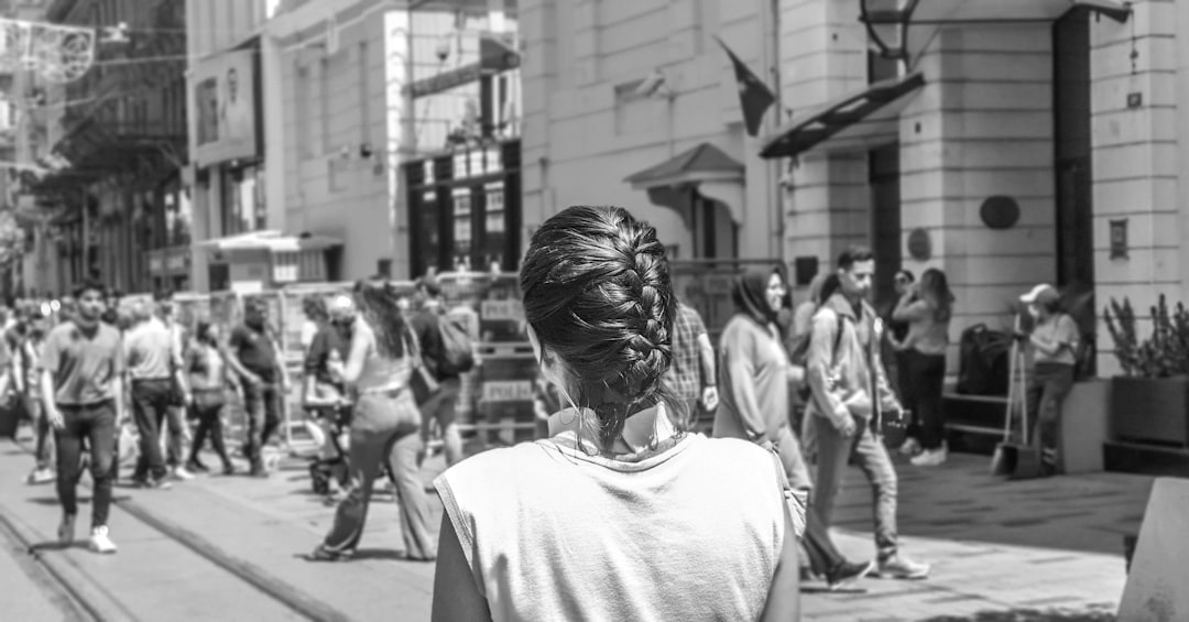 A young woman with her hair in an elegant French braid stands on the street of Milan, surrounded by people who walk along it and look at her. The photo is in the style of black and white street photography, taken from behind the subject with a back view using a Canon EOS R5 camera. –ar 128:67