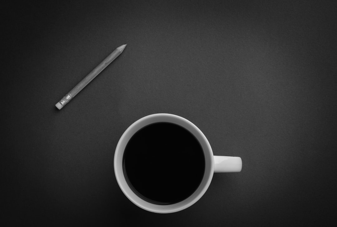 A black and white photo of coffee in a mug with a pencil on the table, in a minimalistic style, with high resolution and great detail, high contrast, high quality photography with a high dynamic range, presented as a flat lay with a top down view and overhead shot, taken with a wide angle lens with a depth of field and macro focus, under studio lighting with a centered composition against a simple background, lit with a high key light creating soft shadows. –ar 64:43