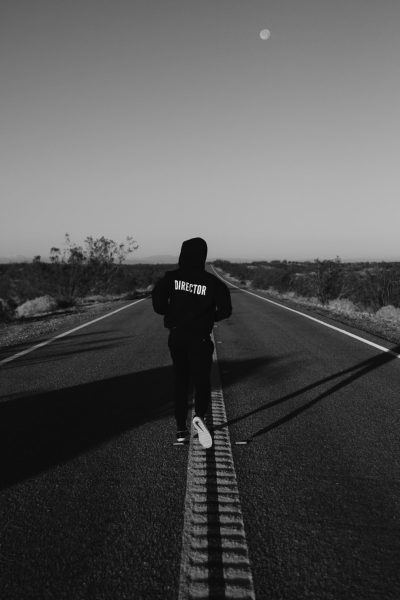 A black and white photo depicts an empty desert road with the moon in view, centered is a man wearing streetwear walking towards the camera with his back to the viewer. He has short hair and wears sneakers. On his hoodie is written "kalREE SDKPUVote." The composition captures him from behind in the style of a cinematic feel with high contrast lighting and a shallow depth of field. --ar 85:128