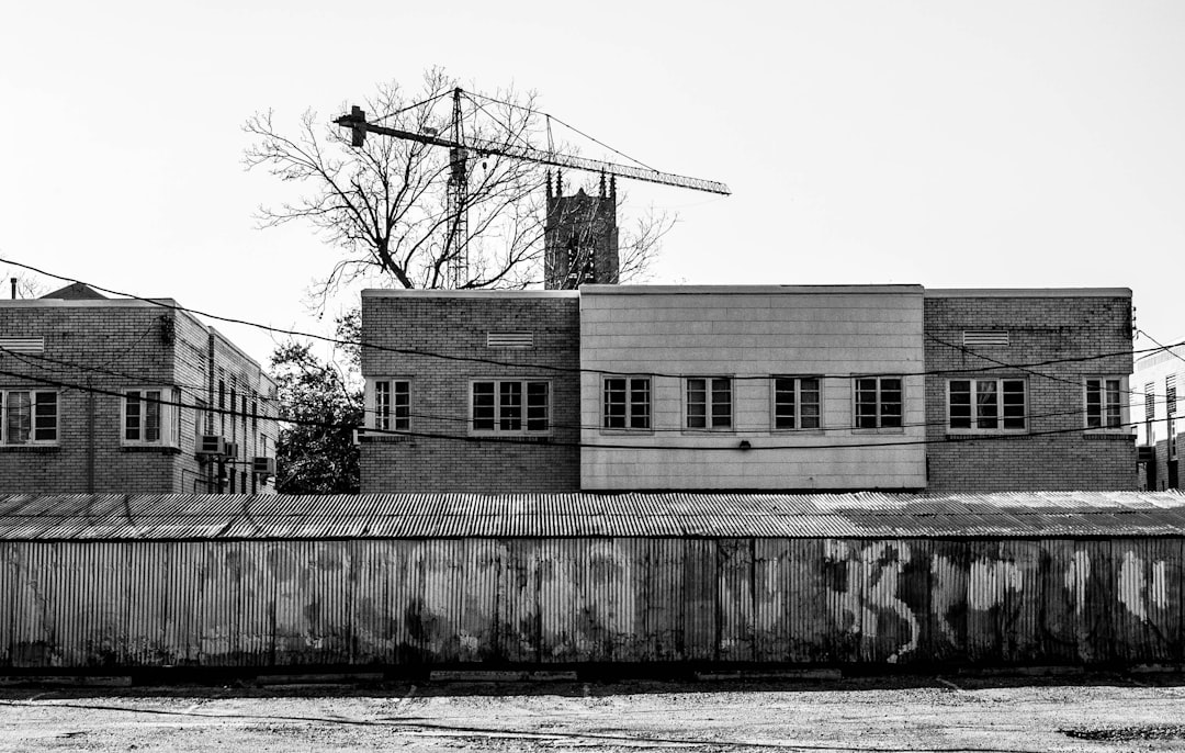 black and white photography, exterior of an industrial building with brick walls in the style of art deco, industrial crane, metal roof, large windows, weathered wood fence, street view, –ar 128:81