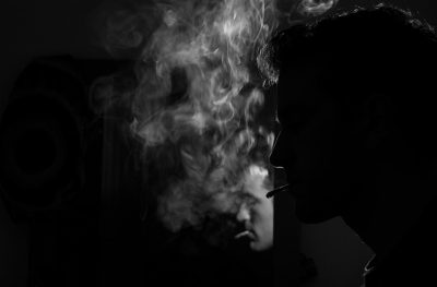 A person smoking, with smoke rising from their mouth and nose against the background of black walls. The scene is captured in the style of silhouette using high contrast photography techniques. High resolution photography. This photo was taken with a Sony Alpha A7 III camera with an f/8 aperture setting. Black & white. --ar 32:21