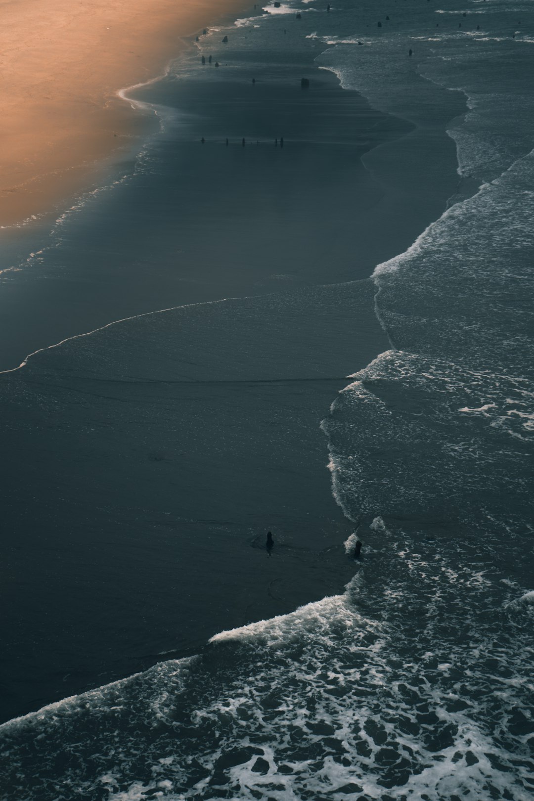 Aerial view of a surfer on the beach, with waves and people in the distance, during sunset, with dark grey tones creating a moody, cinematic style. –ar 85:128