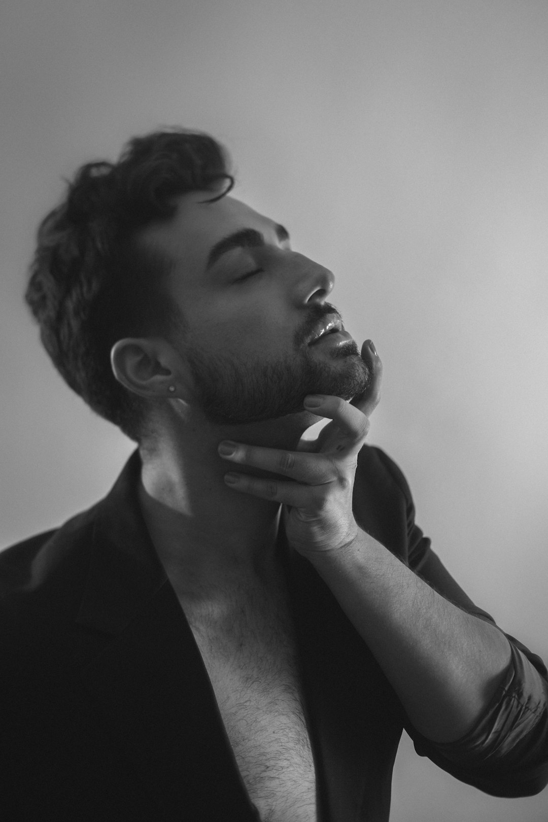 A black and white portrait of an attractive man in his mid-20s with dark hair and a short beard, touching his neck while wearing a blazer against a clean background, in the style of fashion photography and editorial photography with soft light and a cinematic style. –ar 85:128