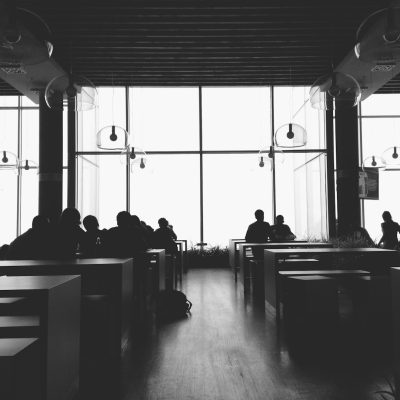 black and white photo of people sitting in a modern interior cafe, large windows in the background, minimalistic style, silhouettes, low angle shot, wideangle lens, natural daylight, soft shadows, minimal elements. The photo has a style similar to that of modern interior photographers with its use of silhouettes, wide angle lens, and natural light creating soft shadows throughout a minimalist setting.