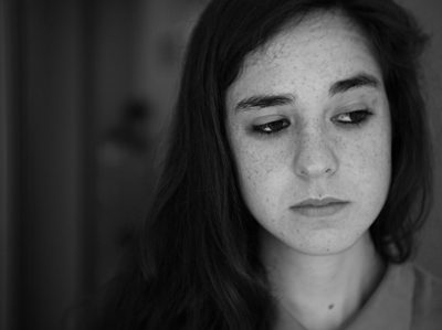 Black and white portrait of sad young woman with long hair, shot on Leica M6 using natural light to capture the subject's expression and features in sharp focus. The background is blurred to emphasize her face. She has dark eyes and freckles across one eye. Her skin looks smooth yet slightly textured due to the film grain effect. --ar 128:95