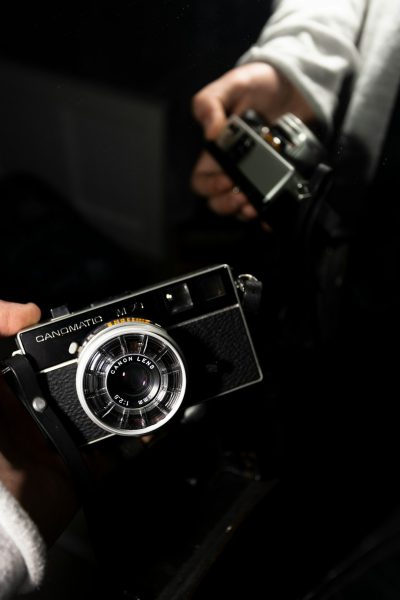 A close up of someone holding an old camera, the person is wearing white and black , the background is dark with some light coming from above, the text "GANYSMagnatoCdessus oAssetto" on top in large letters, the photo was taken in the style of Canon EOS, 50mm portrait lens. --ar 85:128