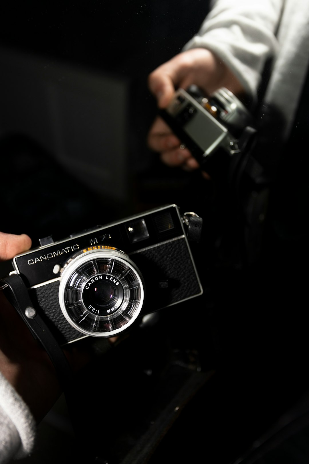 A close up of someone holding an old camera, the person is wearing white and black , the background is dark with some light coming from above, the text “GANYSMagnatoCdessus oAssetto” on top in large letters, the photo was taken in the style of Canon EOS, 50mm portrait lens. –ar 85:128